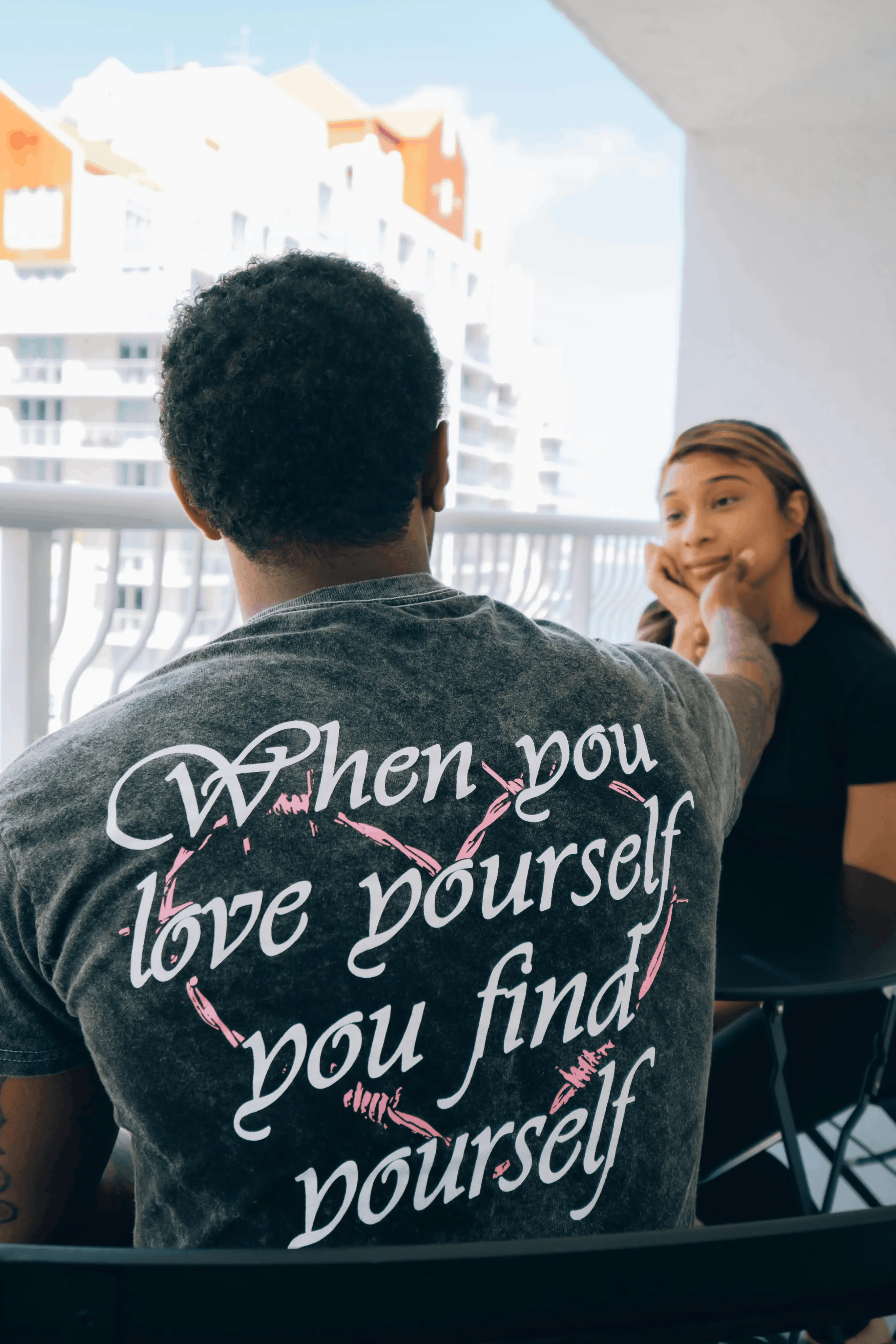 A couple talking on a balcony — one wearing a “When you love yourself you find yourself” shirt